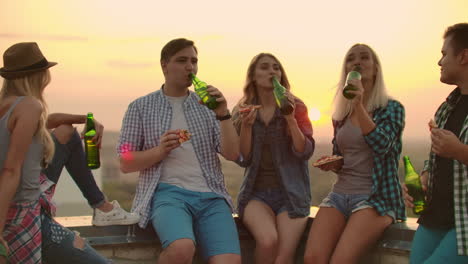 Three-girls-and-three-boys-in-plaid-shirts-drinks-beer-from-green-bottels-on-the-party-with-friends-on-the-roof-at-the-sunset.They-are-sitting-together-eat-hot-pizza-after-in-summer-evening.